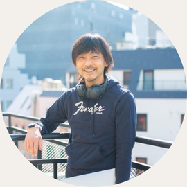Smiling man leaning on a railing on a rooftop, wearing a navy hoodie and headphones around his neck, sunny urban background.